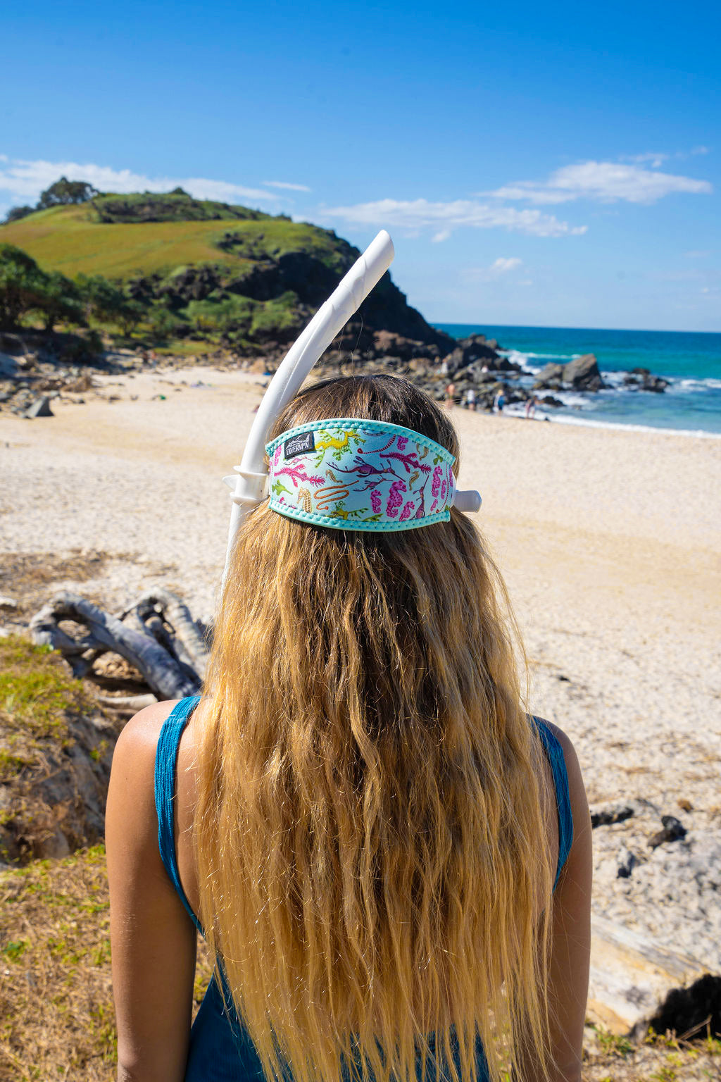 Blonde haired woman seen close up from behind at the beach with the seahorse green design neoprene mask strap tamer fitted to mask ocean illustration by Diver Mon Art 