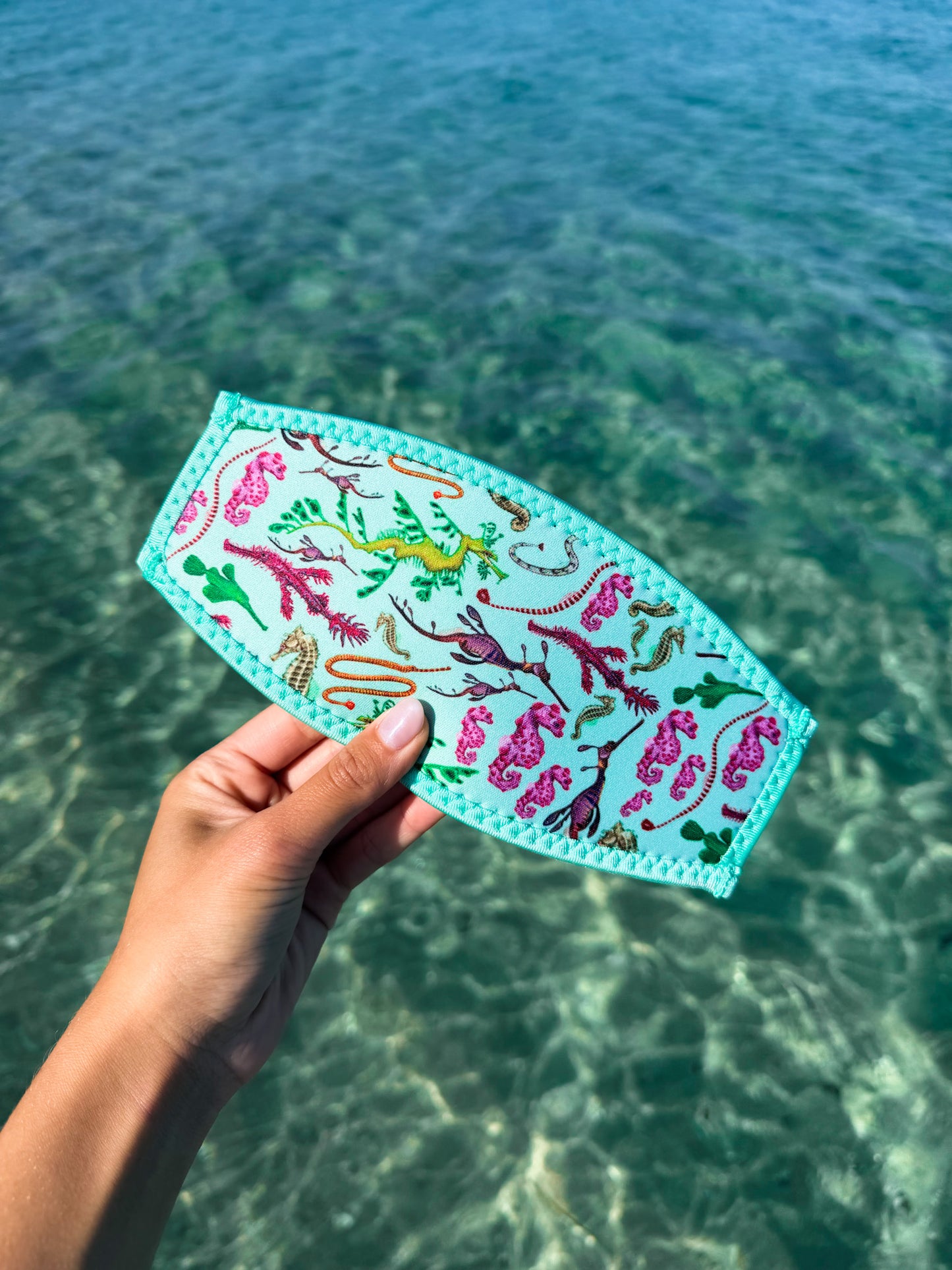 Hand holding seahorse green neoprene mask tamer design by Diver Mon Art design against a clear blue water background