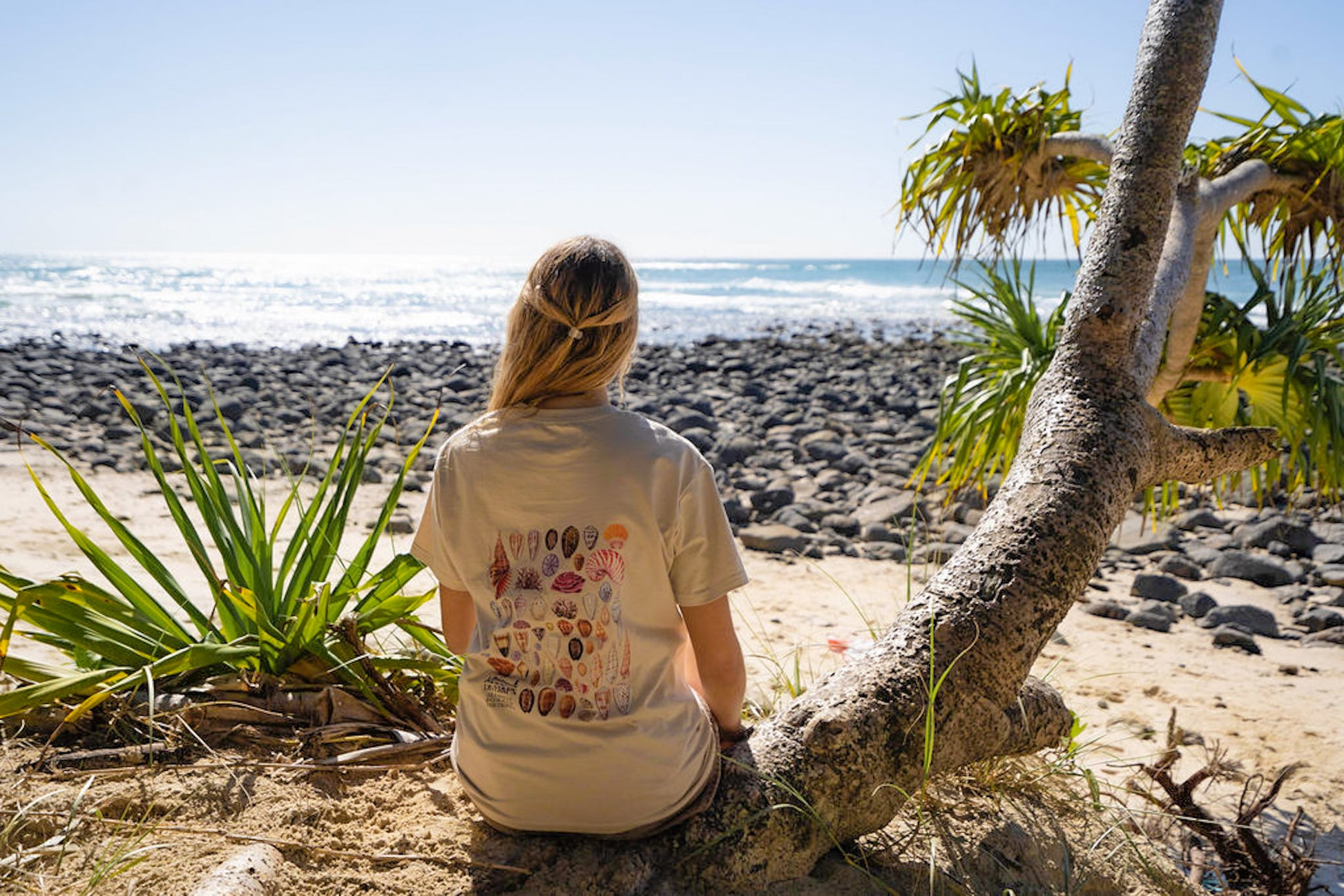 Woman wearing Diver Mon Art shell tee sitting on a beach with a scenic view of the ocean and rocks.