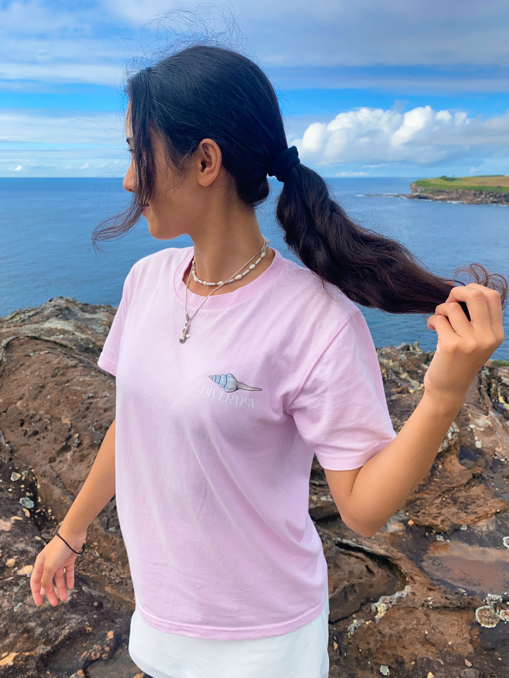 Diver Mon in a pink shell illustration shirt standing on a rocky coastal area with ocean and sky in the background