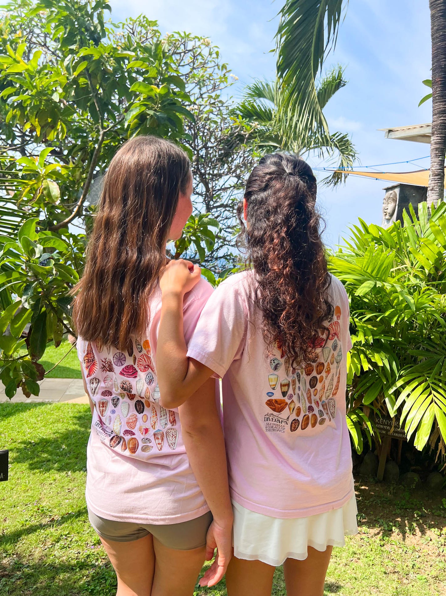 Two women standing back-to-back wearing a Diver Mon Art shell shirt in a garden with greenery and a clear sky.