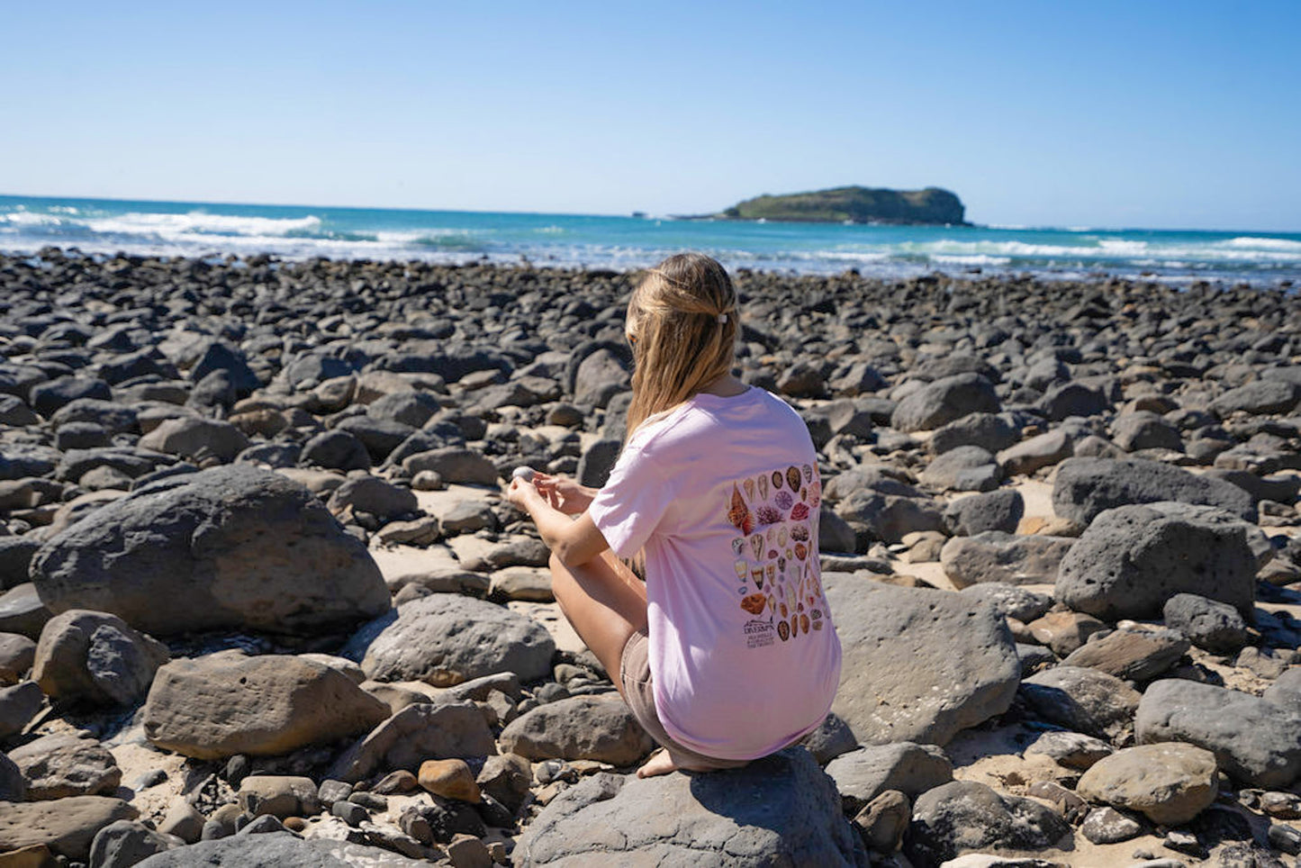 Woman sitting on a rocky beach wearing Diver Mon Art shells with ocean and island in the background