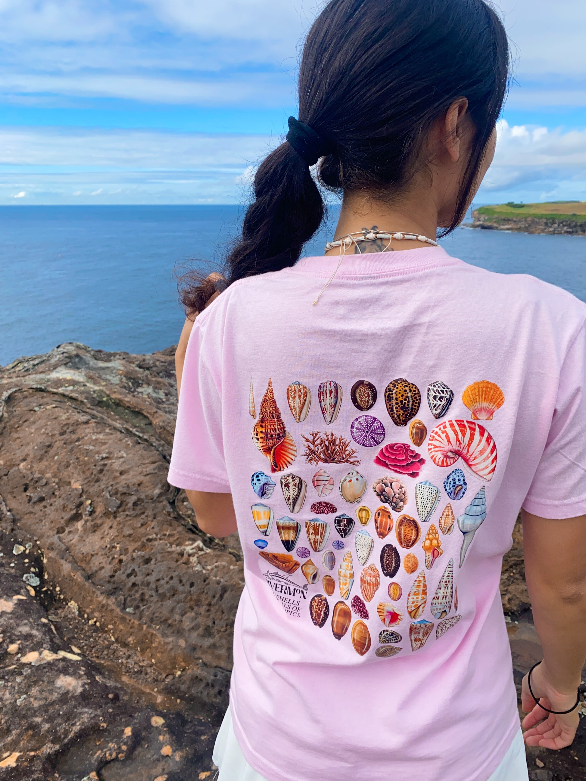 Diver Mon wearing a pink t-shirt with shell designs by the ocean