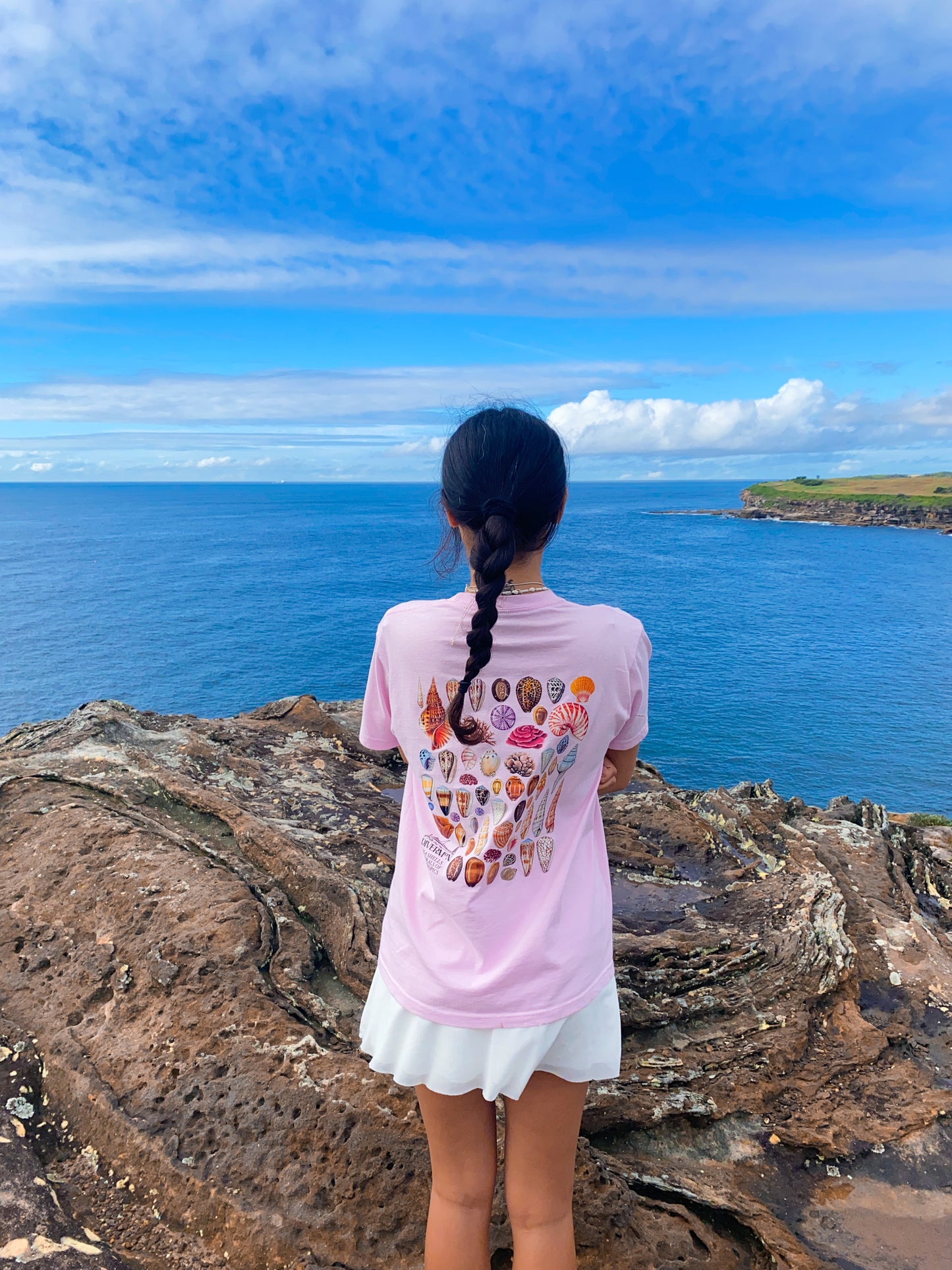 Diver Mon wearing the pink shell shirt with colorful shell designs, standing on a rocky cliff overlooking the ocean. Marine illustrations
