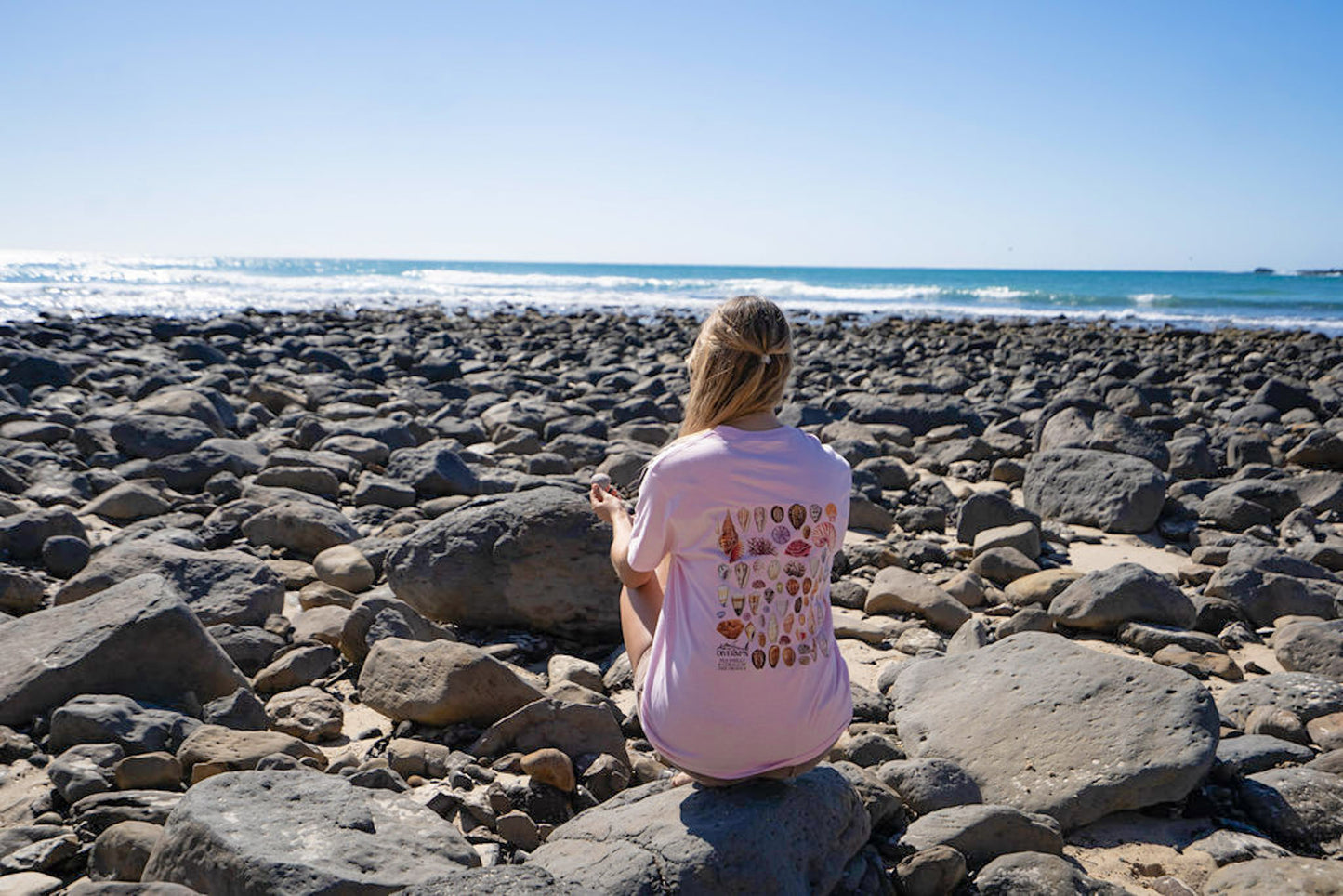 Woman sitting on rocks by the ocean wearing a pink shell shirt by Diver Mon Art with a colorful design.