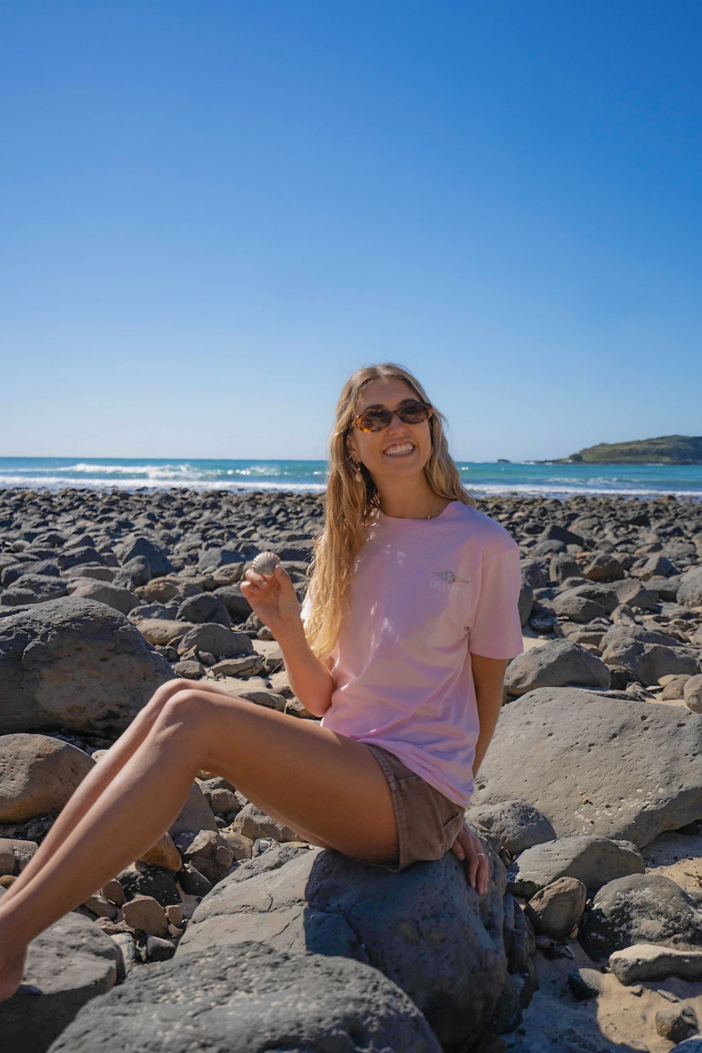 Woman sitting on rocks by the ocean wearing pink shell top by Diver Mon Art with sunglasses on