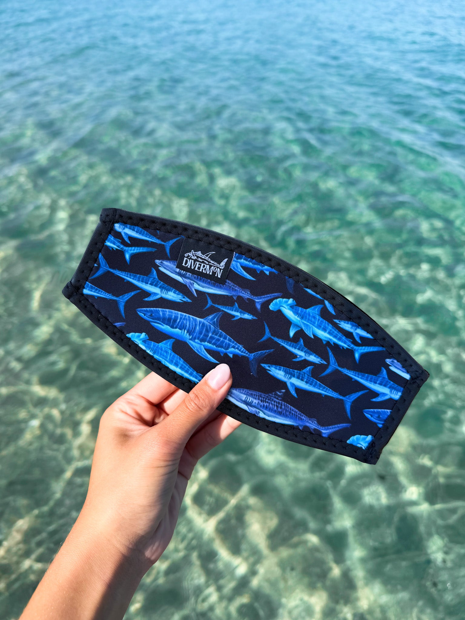 Hand holding a black shark neoprene mask tamer by Diver Mon Art design against a clear blue water background