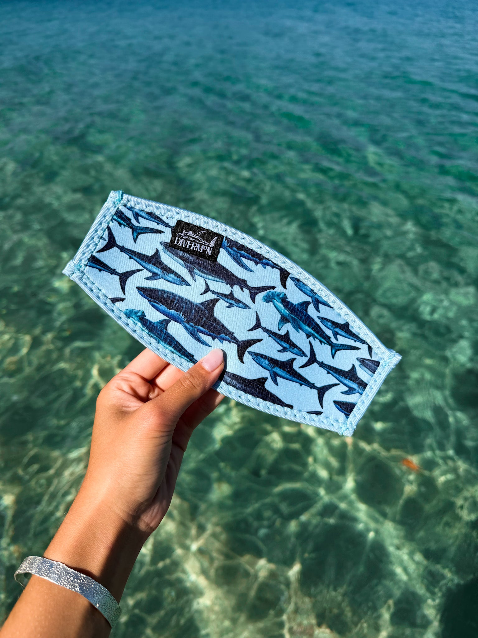 Hand holding light blue shark neoprene mask tamer by Diver Mon Art design against a clear blue water background