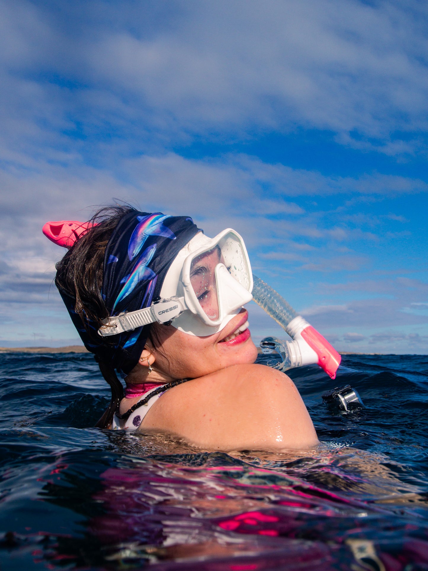 Dark haired woman shown from side view wearing the limited addition thresher black UV gaiter buff design above clear blue water made from recycled polyester-spandex ocean illustration by Diver Mon Art 