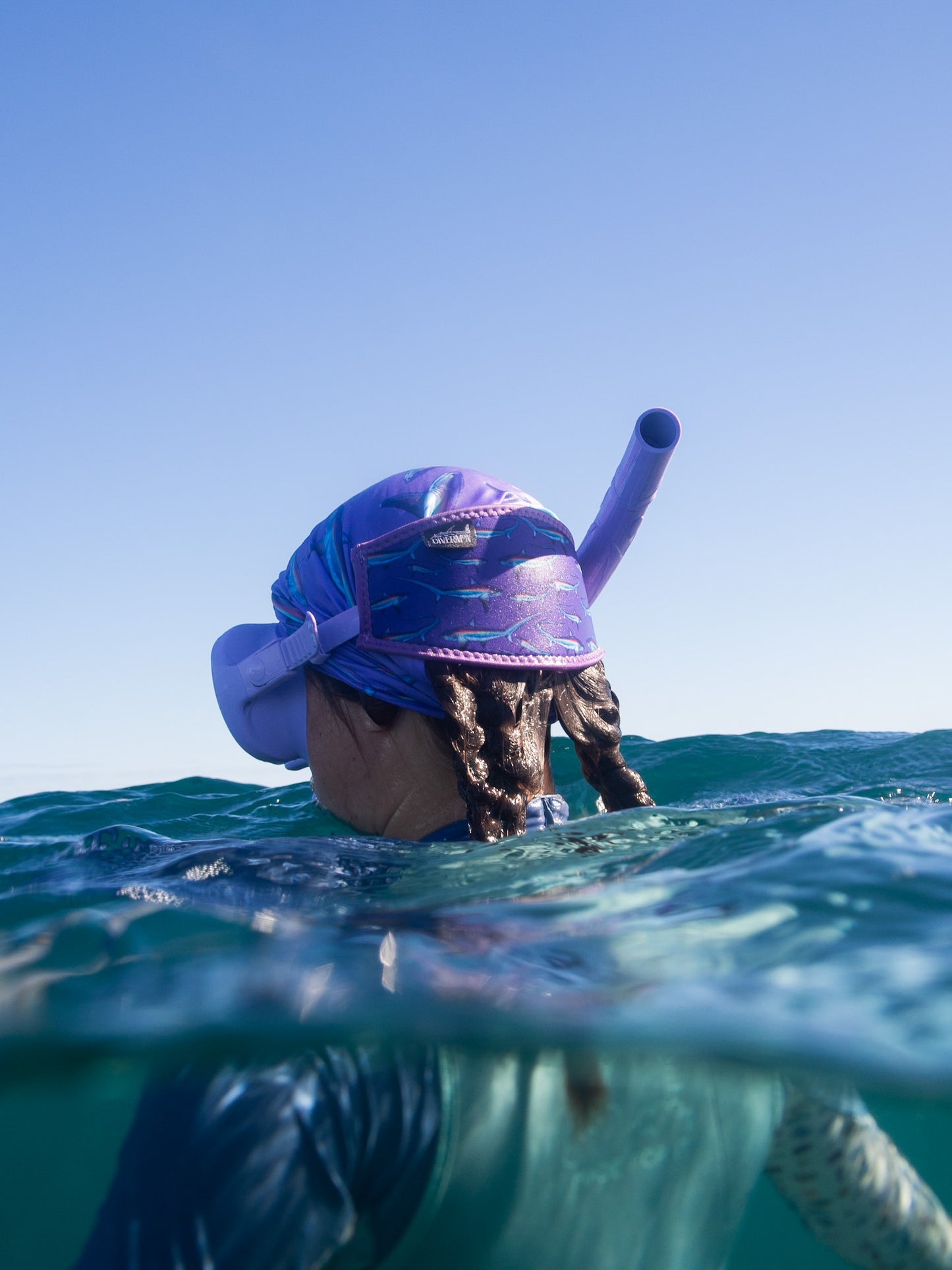 Dark haired woman seen above clear blue water wearing a purple mask fitted with the limited addition purple thresher neoprene mask tamer matching with the purple thresher UV gaiter ocean illustration by Diver Mon Art 