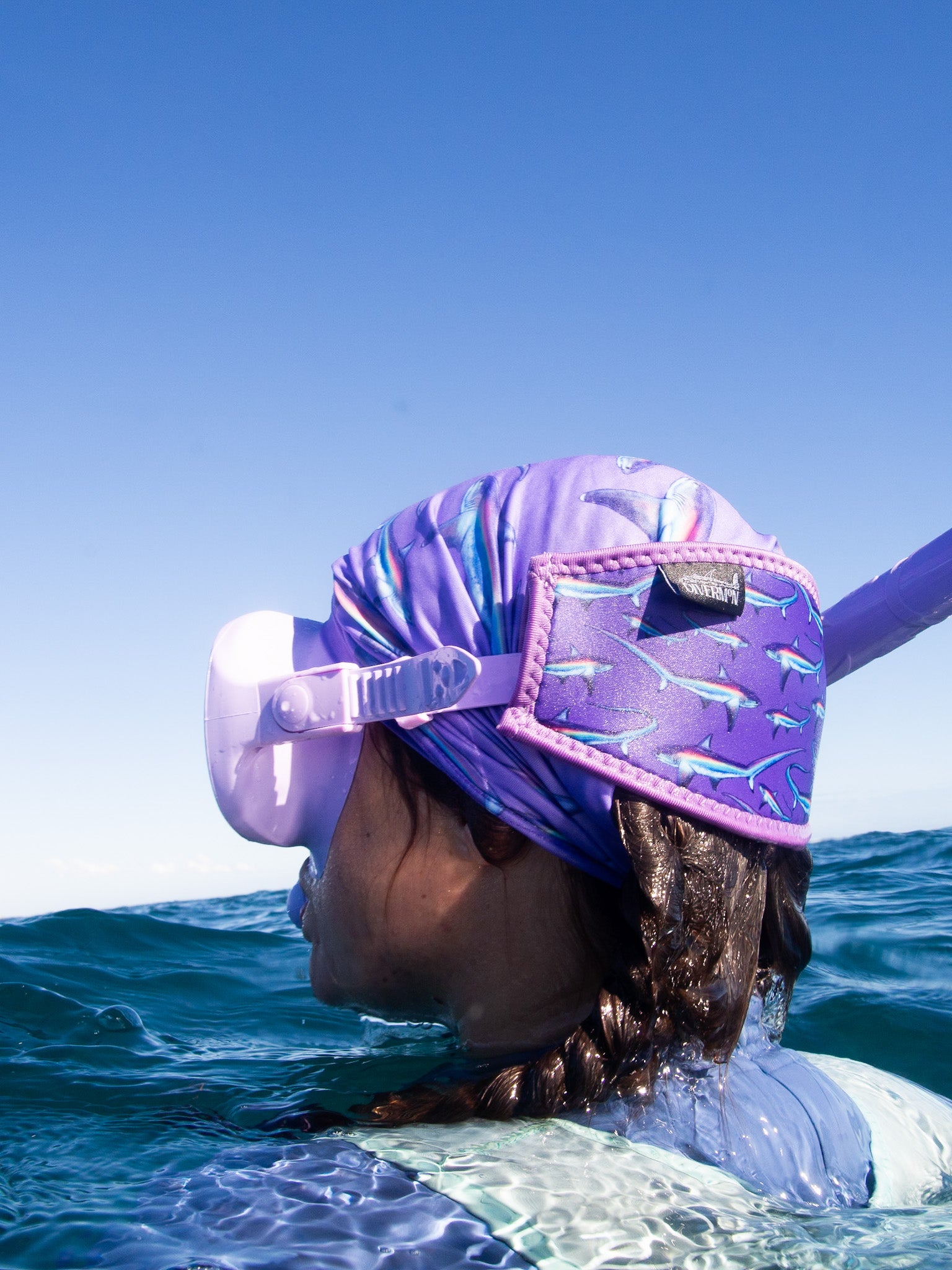 Dark haired woman seen close up side view in great detail above clear blue water wearing a purple mask fitted with the limited addition purple thresher neoprene mask tamer matching with the purple thresher UV gaiter ocean illustration by Diver Mon Art 
