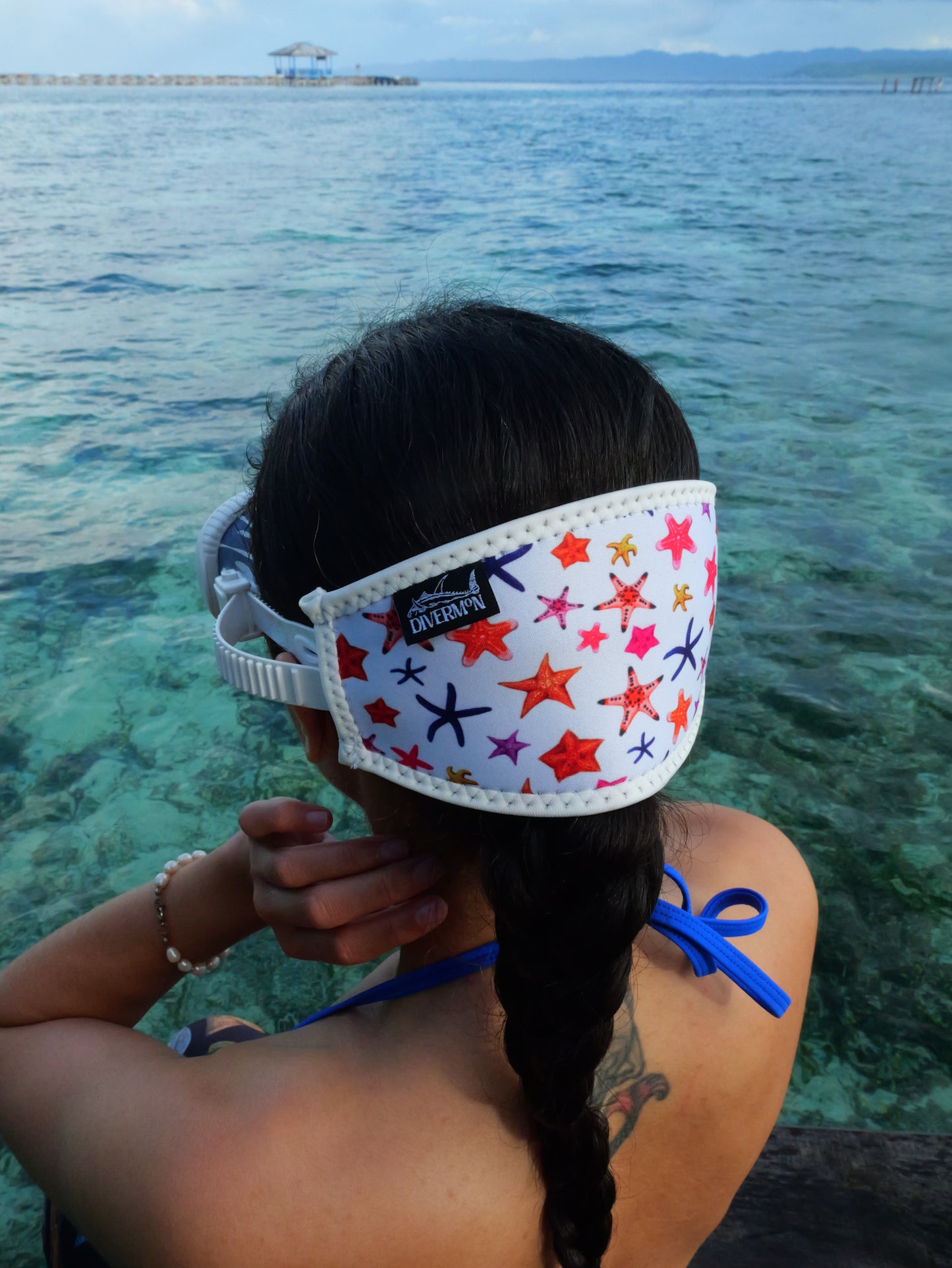 Dark haired woman shown from behind in blue clear water close up wearing a mask fitted with white sea star design neoprene mask strap tamer ocean illustration by Diver Mon Art