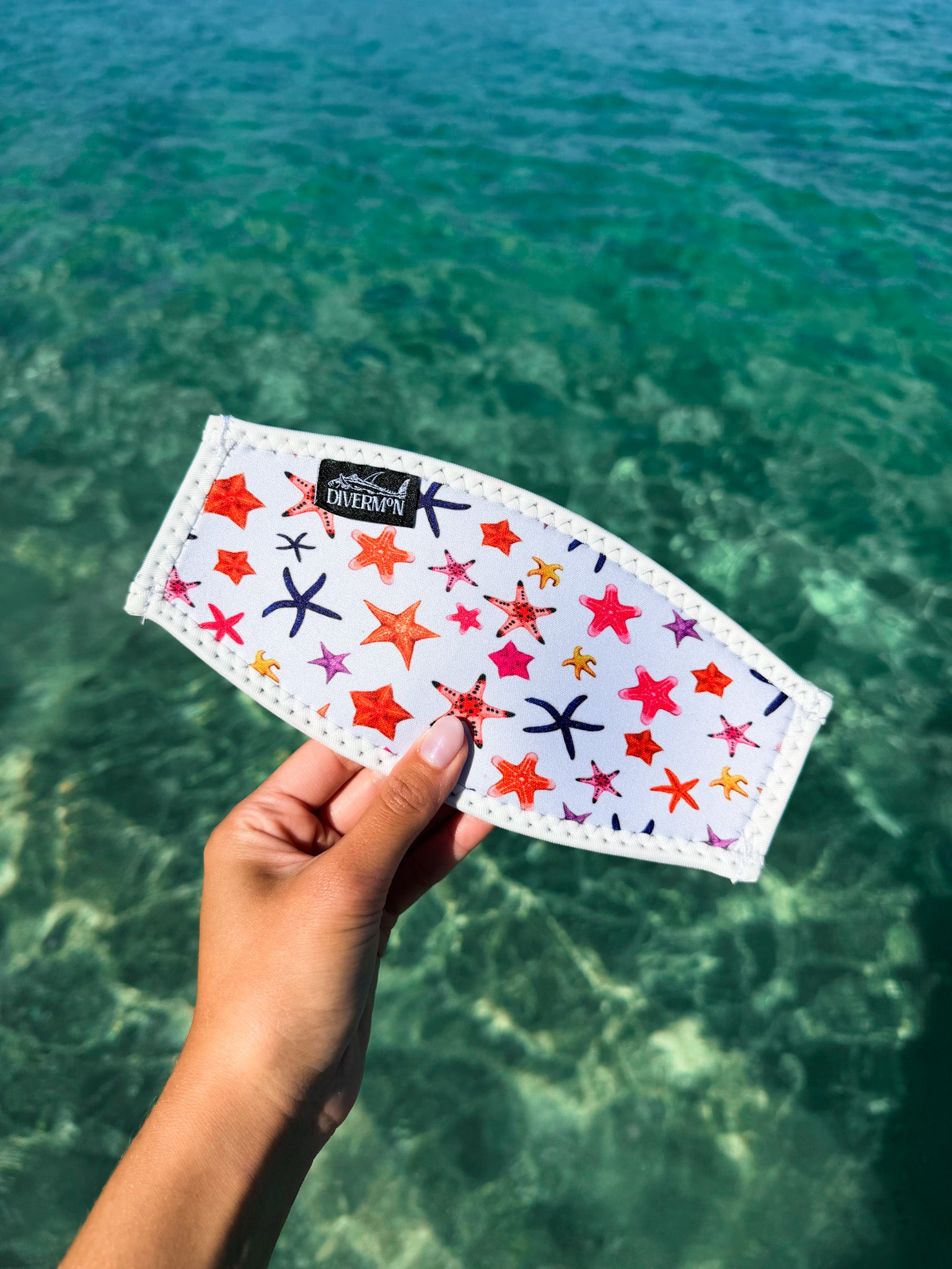 Hand holding the white sea star neoprene mask tamer design by Diver Mon Art design against a clear blue water background