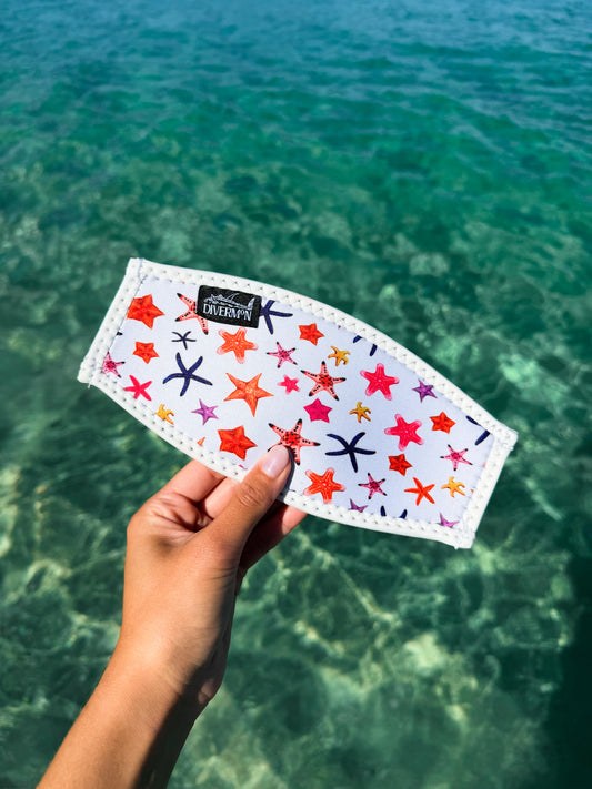 Hand holding the white sea star neoprene mask tamer design by Diver Mon Art design against a clear blue water background
